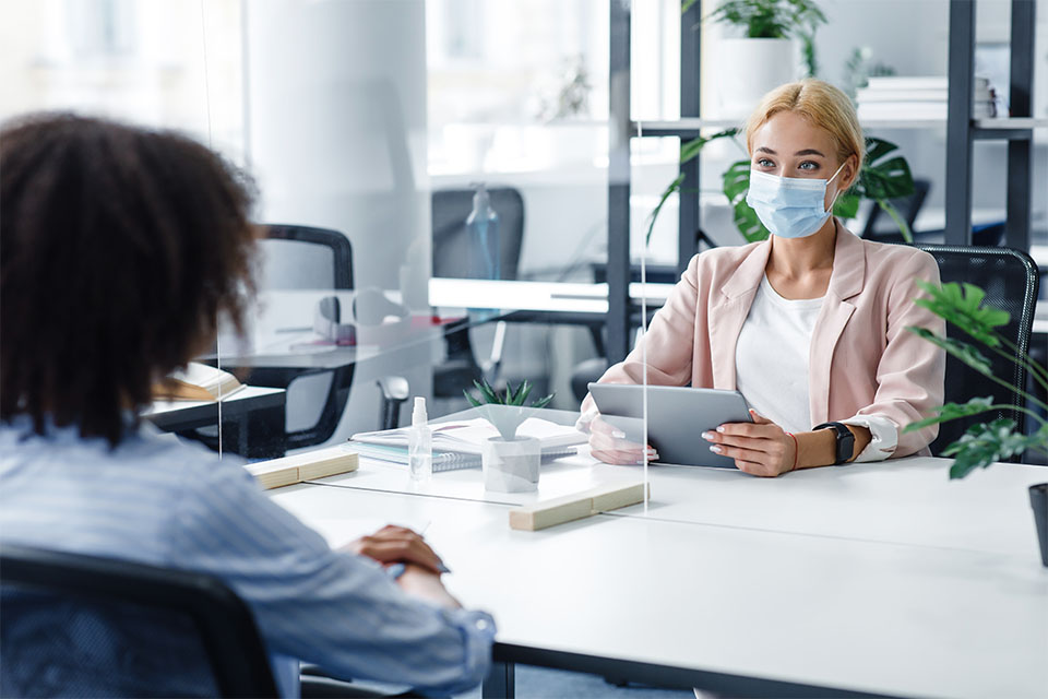 Women talk at desk with protective separator.